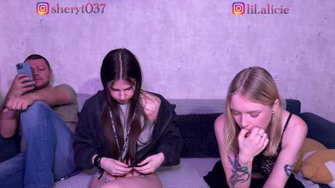 Video of three young women, one with long hair, one with blonde hair, and another with short hair, sitting on a sofa, focused on a smartphone, in a dimly lit room.