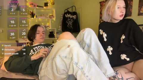Video of two young women, one with short dark hair and the other with platinum blonde hair, lounging on a bed, wearing casual, oversized clothing. Background features a cozy, dimly lit room with shelves and a black bag.
