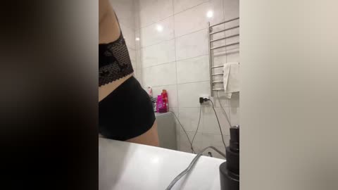 Video of a woman in black lace bra and shorts, standing in a modern, white-tiled bathroom with a silver towel rack and pink shampoo bottle on the sink.