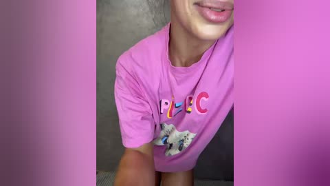 Video of a smiling person wearing a pink T-shirt with \"FEC\" and a cat design, partially obscured by pink frames. Background shows a gray textured wall.