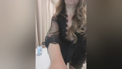A video of a fair-skinned woman with long, wavy blonde hair, wearing a sheer black lace robe, standing in a bathroom. She holds a blue toiletry bottle in her right hand. The background features beige walls and a white bathtub.