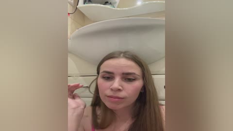 Video of a young woman with long brown hair, wearing a pink tank top, standing in a bathroom with beige walls and a white sink. She is playfully pulling her hair.