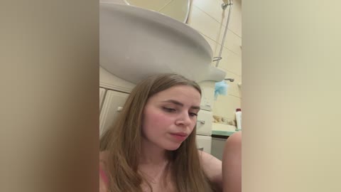 Video of a young woman with long blonde hair, fair skin, and medium build, standing in a bathroom. She has a neutral expression. Background shows a white sink, beige tiles, and toiletries.