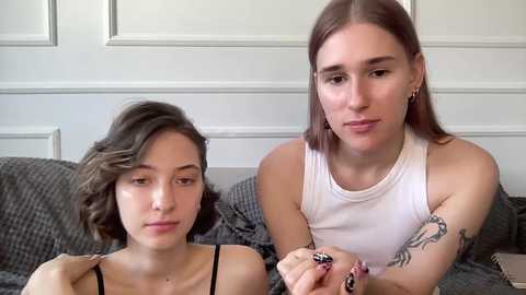 Video of two young women with short hair, one in a white tank top and another in a black top, sitting on a gray quilted couch in a minimalist room with white paneled walls.