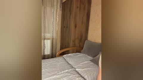 Video of a cozy bedroom corner with a wooden headboard, beige walls, and a gray-patterned quilted bedspread. A wooden chair and sheer curtains with a floral pattern add warmth.