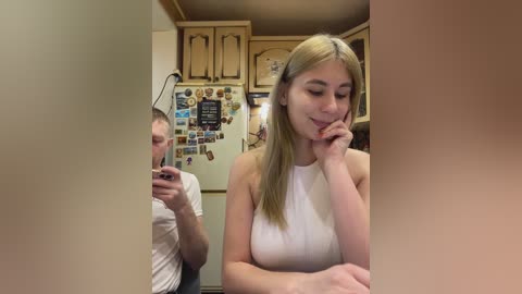Video of a blonde woman in a white tank top, leaning on a kitchen counter, with a man taking a selfie in the background.