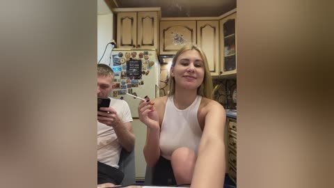 Video of a blonde woman in a white tank top, seated in a kitchen with wooden cabinets, holding a smartphone, and a man in a white t-shirt, both smiling.