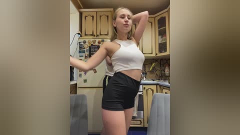 Video of a blonde Caucasian woman with fair skin, wearing a white crop top and black shorts, striking a pose in a cozy kitchen with wooden cabinets, a fridge, and utensils.