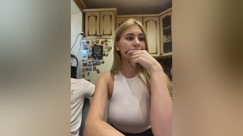 Video of a blonde, fair-skinned woman with medium build, wearing a white tank top, biting her finger while seated in a cozy kitchen with wooden cabinets and a refrigerator covered in magnets.
