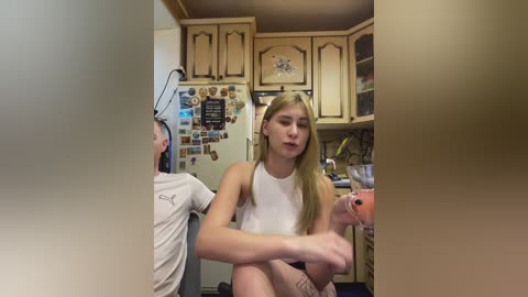 Video of a young woman with long blonde hair in a white tank top, sitting on a kitchen counter, holding a pink cup, surrounded by wooden cabinets, a refrigerator, and a boy in a white shirt.