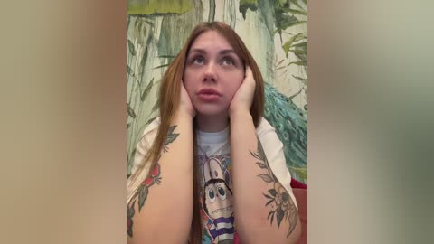 Video of a young woman with long, straight red hair, fair skin, and numerous tattoos on her arms. She wears a white T-shirt with a cartoon character. Background features a green, leafy mural.