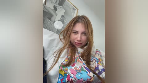 A video of a young woman with long, straight light brown hair, wearing a colorful, cartoon-print robe, standing in front of a black-and-white framed photo on a white wall.