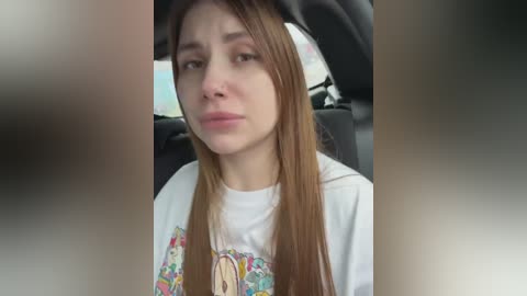 Video of a young woman with long brown hair, wearing a colorful graphic T-shirt, sitting in a car with a neutral expression.