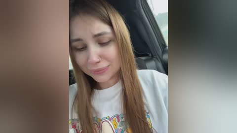Video of a young woman with long brown hair, wearing a colorful graphic t-shirt, seated in a car, eyes closed, looking relaxed.