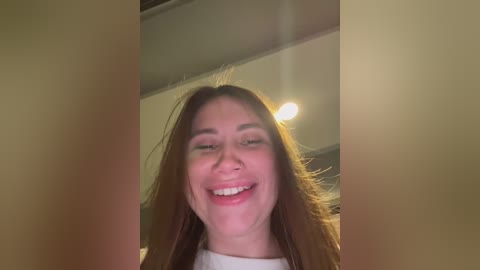 Video of a smiling young woman with long, straight brown hair, wearing a white shirt, standing indoors with a beige wall and ceiling light in the background.