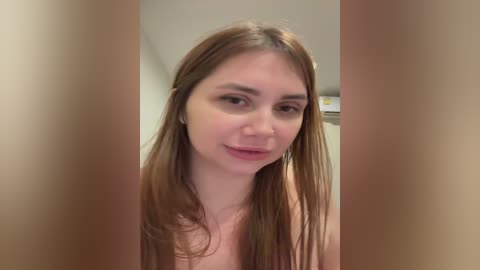 A video of a young Caucasian woman with long, straight brown hair, light skin, and a slender build, standing in a bathroom with beige walls. She has a neutral expression and is not wearing any clothing.