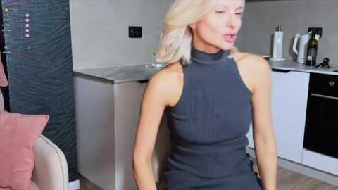 Video of a blonde woman with fair skin in a gray ribbed sleeveless dress, sitting in a modern kitchen with stainless steel appliances and a pink cushion.