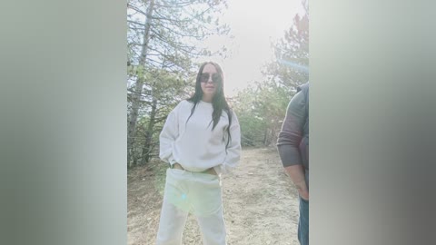 Video of a woman with long dark hair, wearing a white hoodie and pants, standing confidently in a forest. The background features sparse trees and a dirt path.