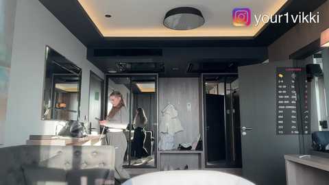 Video of a modern hotel room with a sleek, minimalist design. A woman with light skin and long brown hair stands near a mirrored vanity, wearing a brown jacket. The room features dark wood and gray tones, with a round ceiling light and a visible time display on the wall.