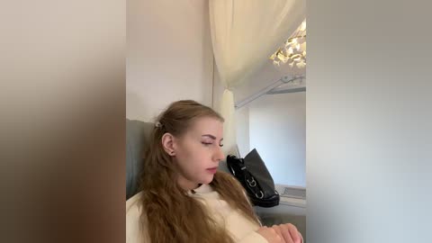 Video of a young Caucasian woman with long, wavy blonde hair, wearing a white top, sitting on a white chair, in a room with beige walls and a chandelier.