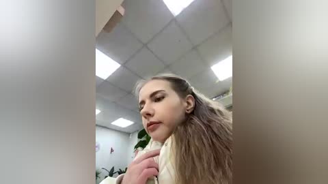 Video of a young woman with long, straight, light brown hair, wearing a beige sweater, sitting in a room with white walls, a ceiling with fluorescent lights, and green plants in the background.