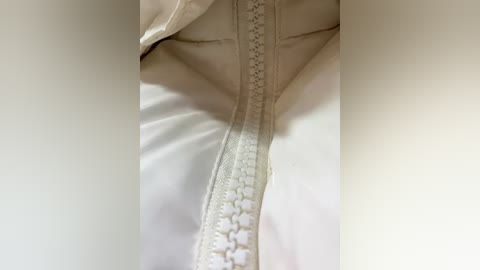 A close-up video of a white zipper running vertically down the center of a white jacket, with the fabric slightly textured and the zipper\u2019s teeth clearly visible. The image is cropped tightly, focusing on the zipper and the jacket\u2019s material.