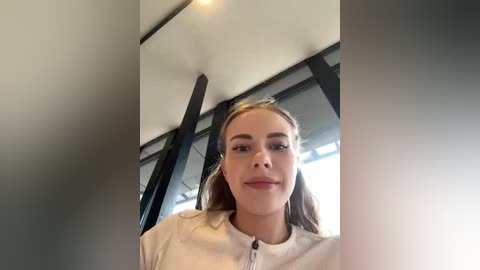 Video of a young Caucasian woman with light skin, straight brown hair, and fair complexion, wearing a white top, indoors with modern architectural elements like large windows and black beams.