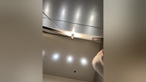 Video of a modern ceiling with metallic, curved surfaces, featuring three recessed, circular lights and a hanging fixture. The image is blurred, focusing on the ceiling and a hand partially obscuring the view.
