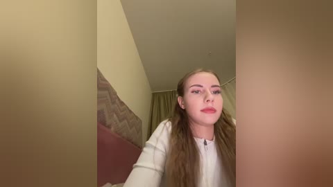 A video of a young Caucasian woman with long, straight brown hair, fair skin, and red lipstick, wearing a white top, standing indoors with beige walls and a patterned headboard in the background.