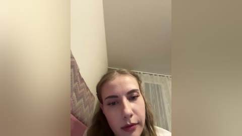 Video of a young woman with light skin, long brown hair, and slight smile, standing in a dimly lit room with beige walls and a partially visible brick wall.