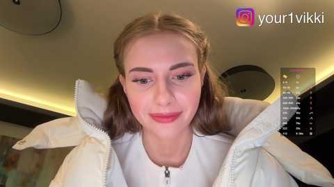 Video of a smiling Caucasian woman with light brown hair styled in pigtails, wearing a white jacket, indoors with a modern, dimly lit background.