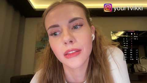 Video of a young Caucasian woman with light skin, long blonde hair, wearing white headphones, smiling, indoors. Background features a dimly lit room with a flat-screen TV.