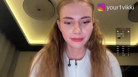 Video of a fair-skinned young woman with long, wavy blonde hair styled in pigtails, wearing a white top, smiling slightly. Background features a modern, dimly lit room with recessed lights and a digital display.