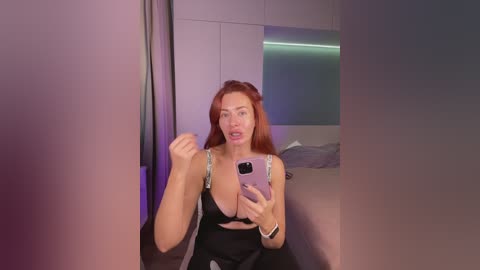 Video of a fair-skinned, red-haired woman in a black bra, taking a selfie in a modern bedroom with purple lighting.