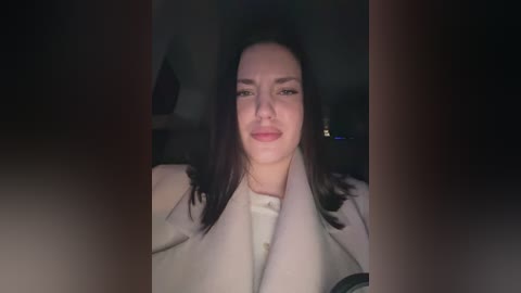 Video of a light-skinned woman with straight, shoulder-length dark hair, wearing a white coat, seated in a dimly lit car.
