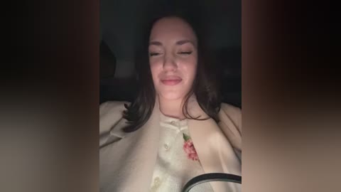 A video shows a smiling woman with dark hair, wearing a beige jacket with floral embroidery, seated in a dimly lit car.