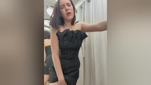 Video of a fair-skinned woman with dark hair, wearing a black, ruffled strapless dress, standing in a bathroom with white curtains and a mirror. She appears distressed, eyes closed, mouth slightly open.