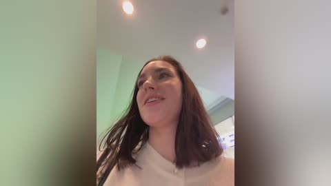 Video of a smiling, light-skinned woman with long, dark hair, wearing a white top, standing indoors near a white wall with recessed lights.