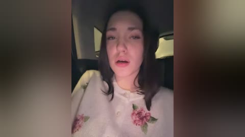 Video of a woman with fair skin and dark hair, wearing a white blouse adorned with floral embroidery, sitting in a dimly lit car, mouth slightly open as if speaking.