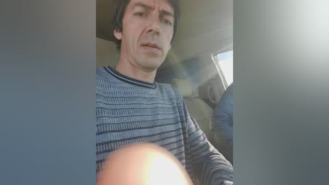 Video of a man in a blue striped sweater, looking serious, sitting in a car with sunlight streaming through the window, creating lens flare.