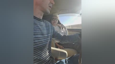 Video of a man in a striped sweater driving a car, with a woman in glasses and a white blouse in the passenger seat, sunlit sky visible through the window.