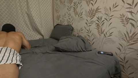 Video of a man lying face down on a bed with gray sheets and pillow, wearing striped boxer shorts, in a room with patterned wallpaper and a television remote on the nightstand.
