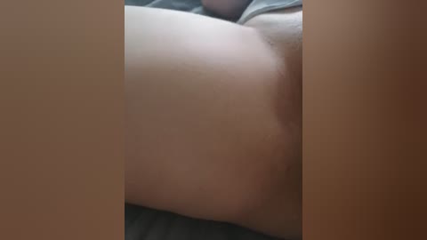 Video of a close-up view of a light-skinned person's bare buttocks, lying on a grey fabric surface, possibly a bed or couch. The image focuses on the smooth, slightly rounded contours of the skin, with a blurred background.