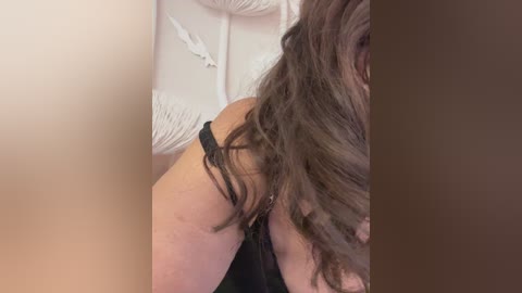 Video of a woman with long, wavy brown hair, partially covering her face. She is wearing a black lace bra, showcasing her medium-sized breasts. The background features a white, textured wall. The image is intimate and focused on her upper body.