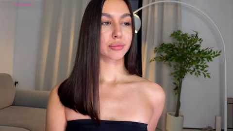 Video of a young woman with straight, dark brown hair, fair skin, and a slender physique, wearing a strapless black top. She stands in a modern living room with beige walls, a tall plant, and a white arch lamp.