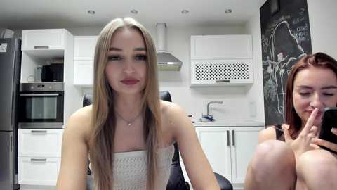 Video of two young women in a modern kitchen. Blonde woman with straight hair, wearing a white strapless top, sits in the foreground. Redhead, topless, with long hair, holds her breasts, smiling in the background. White cabinets, stainless steel appliances, and a chalkboard wall adorn the scene.