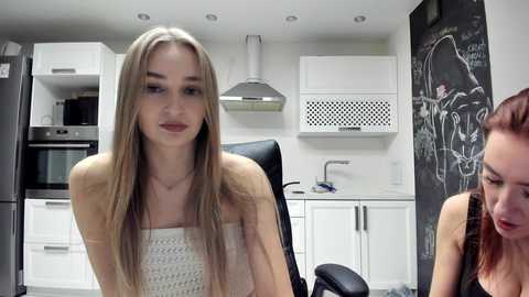 Video of a young, slim, fair-skinned woman with long, straight blonde hair in a white tank top, sitting in a modern, white kitchen with stainless steel appliances and a chalkboard wall.