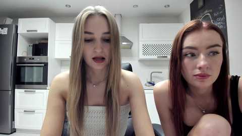 Video of two young women in a modern kitchen: blonde in white top, redhead in black top. White cabinets, stainless steel appliances, and black stools in background.