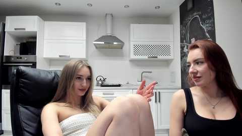 Video of two young women with fair skin, one in a black strapless dress, the other in a white patterned dress, seated in a modern kitchen with white cabinets, stainless steel appliances, and a blackboard.