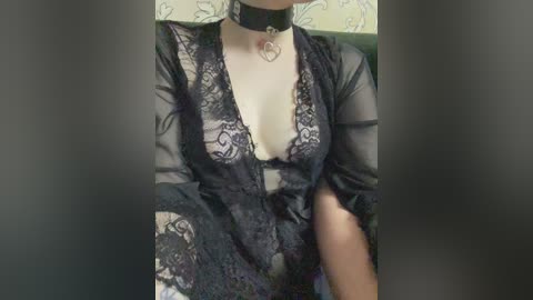 Video of a woman in black lace lingerie and a choker, seated with legs apart, showing cleavage and thigh tattoo.